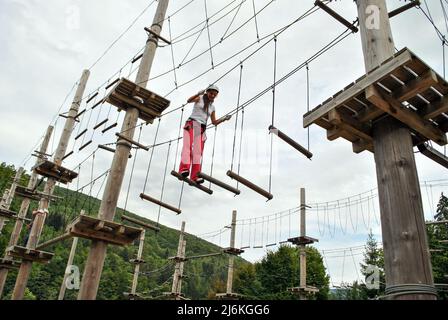 Eine junge Frau in einem Seilpark überquert eine Hängebrücke aus Holz in einem Geschirr Stockfoto