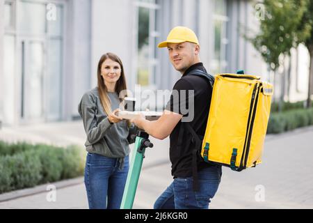 Kurierdienst Arbeiter auf Elektro-Push-Roller liefert Lebensmittel an Kunden, Online-Bestellung Stadt Versand. Junger Mann auf Roller mit gelbem Thermobeutel gibt Pizzakarton an Frau Klientin. Stockfoto