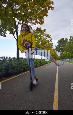 Eine junge Frau fährt perspektivisch mit einem Roller in einer gelben Jacke einen Weg entlang und lacht Stockfoto