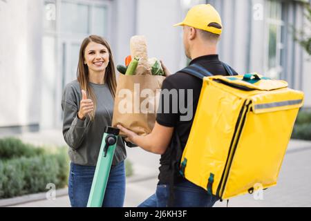 Kurierdienst Arbeiter auf Elektro-Push-Roller liefert Lebensmittel an Kunden, Online-Bestellung Stadt Versand. Junger Mann auf Roller mit Thermobeutel gibt Papiertüte mit frischem Gemüse an Frau Client. Stockfoto