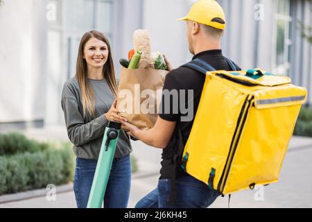 Kurierdienst Arbeiter auf Elektro-Push-Roller liefert Lebensmittel an Kunden, Online-Bestellung Stadt Versand. Junger Mann auf Roller mit Thermobeutel gibt Papiertüte mit frischem Gemüse an Frau Client. Stockfoto