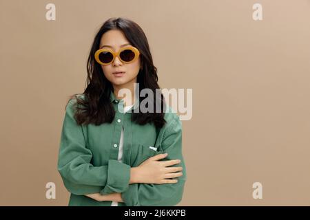 Modische liebenswert koreanische junge Frau in khaki grün Hemd stilvolle Brille Falten Hände posiert isoliert auf über beige Pastell Studio Hintergrund. Cool Stockfoto