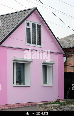 Rosa Fassade eines einstöckigen Hauses mit einem dreieckigen Dach. Kunststofffenster im Haus mit Metalljalousien oben. Das Fenster im Dachgeschoss. Stockfoto