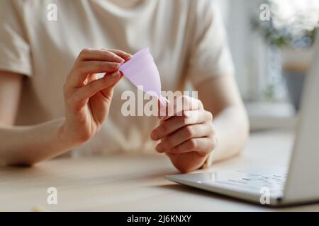 Nahaufnahme einer jungen Frau, die einen rosa Menstruationstasse am Laptop hält und nach weiblichen Produkten forscht Stockfoto