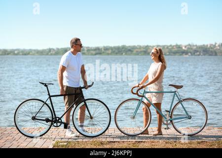 Reifes Paar mit Fahrrädern, das am Sommertag am Flussufer entlang läuft Stockfoto