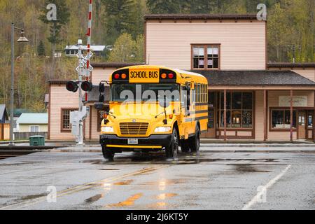 Skykomish, WA, USA - 26. April 2022; Gelber Schulbus über Bahngleise in Skykomish Washington. Die Straße ist nass im Regen. Stockfoto