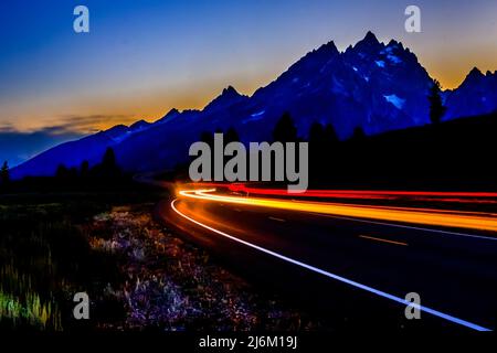 Die Autobahn im Grand Teton National Park, die vom Signal Mountain in Richtung Parkzentrale führt. Sonnenuntergang, Autolampen Streifen um die Kurve. Stockfoto