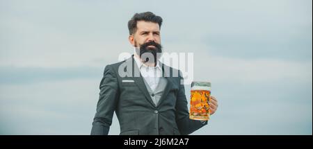 Hipster mit Bart und Schnurrbart im Anzug trinkt Bier. Bierzeit. Ein hübscher Barkeeper hält ein Glas Bier am Himmel. Banner, Kopierbereich. Stockfoto