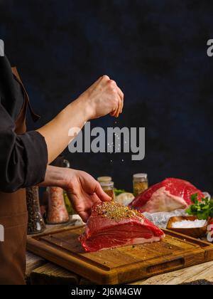 Ein Koch in dunkler Uniform streut Gewürze auf ein großes Stück Fleisch auf dunkelblauem Hintergrund. Gewürze im gefrorenen Flug. Nahaufnahme. Das Konzept ist Kochen Stockfoto