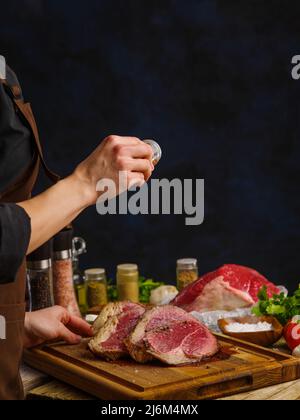 Saftige, rohe Rindersteaks werden von einem professionellen Koch auf dunkelblauem Hintergrund zubereitet. Gemüse, Kräuter, Gewürze, Gewürze auf dem Küchentischen Stockfoto