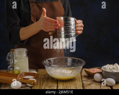 Der Koch siebt Mehl durch ein Sieb auf einem hölzernen Küchentisch. Mehl im gefrorenen Flug. Zutaten für die Teigherstellung auf dunkelblauem Hintergrund. T Stockfoto
