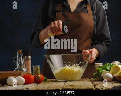 Der Koch fügt geriebenen Käse in einer großen Glasschüssel Gewürze hinzu. Levitation. Gemüse, Kräuter, Eier, Gewürze, Gewürze auf einem Holztisch, dunkelblauer Rücken Stockfoto