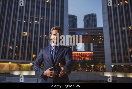 Glücklicher lächelnder Geschäftsmann in Anzug in der Nähe des Büros, erfolgreicher Arbeitgeber, um einen Deal zu machen, während er nachts in der Nähe des Wolkenkratzers steht. Partner wackeln Stockfoto