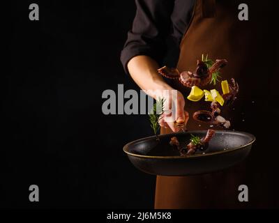 Ein professioneller Koch kocht Oktopus mit Zitronenscheiben in einer Bratpfanne auf schwarzem Hintergrund. Gefrorenes Essen während des Fluges. Restaurant, Hotel, Bankett, Picknick. Stockfoto