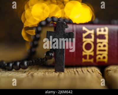 Nahaufnahme. Auf einem Holztisch ist ein rotes Buch - die Heilige Bibel und ein Rosenkranz mit einem Kreuz auf gelbem Hintergrund mit Lichtern. Symbole des Katholizismus, Chris Stockfoto