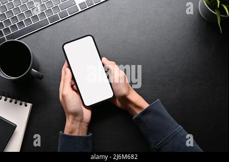 Draufsicht, Ein Mann Hände mit Handy auf seinem dunklen stilvollen Arbeitsplatz-Tisch. Draufsicht, Telefon weißen Bildschirm mockup. Stockfoto