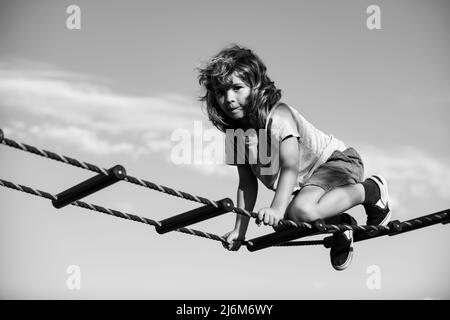 Kind klettert im Netz. Netter Junge klettert die Leiter auf dem Spielplatz hoch. Kind klettert die Leiter gegen den blauen Himmel hinauf. Stockfoto