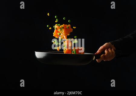 Ein professioneller Koch bereitet verschiedene Meeresfrüchte und Gemüse in einer Pfanne auf schwarzem Hintergrund zu. Gefrorenes Essen während des Fluges. Restaurant, Hotel, Cafe, ho Stockfoto