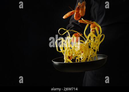 Ein professioneller Koch bereitet italienische Pasta mit Garnelen in einer Pfanne auf schwarzem Hintergrund zu. Gefrorenes Essen während des Fluges. Restaurant, Hotel. Rezepte für RE Stockfoto
