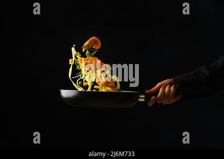 Der Prozess der Zubereitung italienischer Pasta mit Meeresfrüchten und Gemüse in einer Pfanne auf schwarzem Hintergrund von einem professionellen Koch. Levitation. Rezepte für Stockfoto