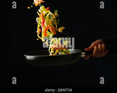 Ein professioneller Koch bereitet Meeresfrüchte mit Gemüsestücken und Kräutern in einer Pfanne zu. Essen auf schwarzem Hintergrund bei einem gefrorenen Flug. Rezepte für r Stockfoto