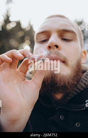 Junger Mann Bart raucht Zigarette oder Tabak. Guy ist süchtig nach Zigaretten und raucht jeden Tag viel Tabak. Draußen raucht er Zigarette Stockfoto