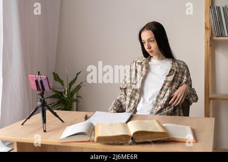 Teenager-Mädchen, das online mit dem Smartphone studiert, Student, der zu Hause mit Schulbüchern am Tisch sitzt, auf einer Videokonferenz das Gespräch hört. Entfernung Stockfoto