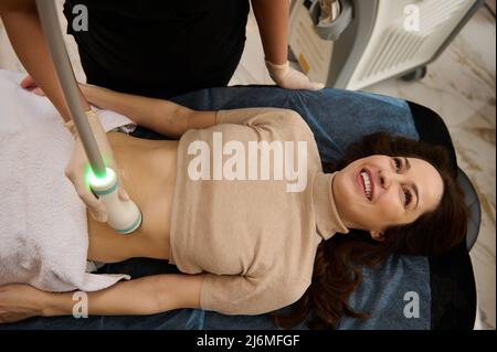 Draufsicht auf eine junge Frau, die in einer Kurklinik eine Mikrostimulation der Bauchmuskeln mit einem speziellen Hochfrequenz-Kavitationssensor erhält. Hardw Stockfoto