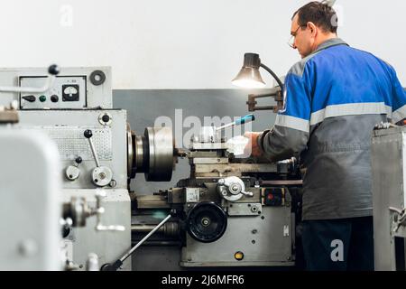 Turner steht hinter der Drehmaschine in der Produktionshalle und arbeitet. Blick auf den Arbeiter von hinten in Overalls. Authentische Arbeitsprozess-Szene in der Produktion. Echter Arbeiter. Nicht inszenierte Szene. Stockfoto