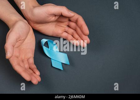 Nahaufnahme von männlichen Händen und blauem Band auf grauer Studiohintergrundwand. Prostatakrebs-Bewusstsein, Movember Men's Health Awareness. Gesundheitswesen, Interna Stockfoto