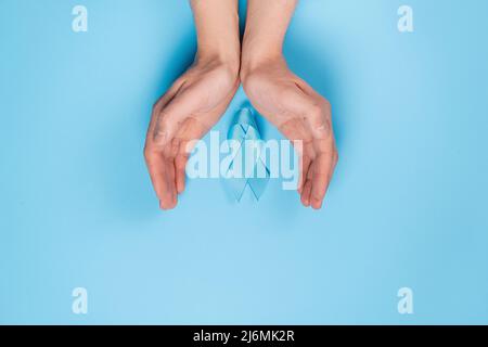 Nahaufnahme von männlichen Händen und blauem Band auf blauer Studiohintergrundwand. Prostatakrebs-Bewusstsein, Movember Men's Health Awareness. Gesundheitswesen, Interna Stockfoto