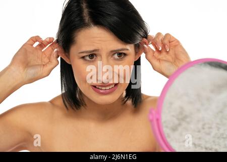 Frau, die sich im Spiegel ansieht und ihre Ohren auf weißem Hintergrund zieht Stockfoto