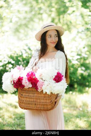 Fröhliches lächelndes Mädchen in einem Strohhut, das einen Korbkorb mit weißen und rosa Pfingstrosen-Blumen in den Straßen einer europäischen Stadt hält. Lifestyle Stockfoto