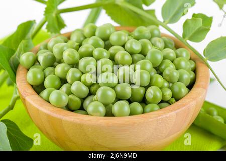 Frische grüne Erbsen in einer Holzschüssel mit Erbsen Pflanzen Blätter auf einer grünen Serviette auf weißem Hintergrund, gesundes Bohnenprotein Stockfoto