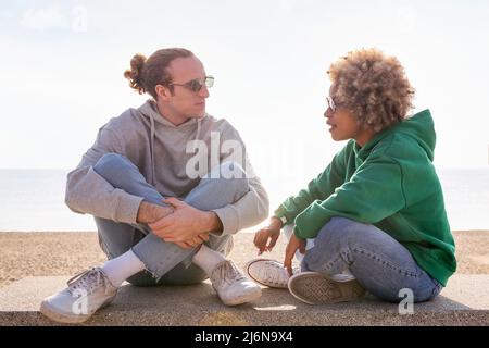 Junges Paar, das beim Sonnenuntergang auf einer Bank am Strand sitzt, Konzept der Freundschaft und Kommunikation in einer Beziehung chattet Stockfoto