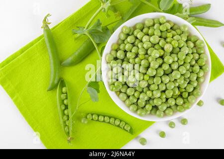 Frische grüne Erbsen in einer Schüssel mit Erbsen Pflanzen Blätter auf Serviette auf weißem Hintergrund, gesundes Bohnenprotein, Draufsicht Stockfoto