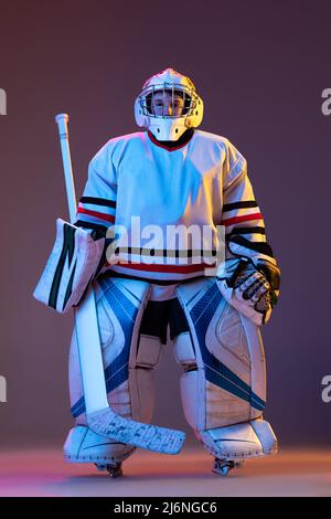 Porträt eines Jungen, Kind posiert in schützender Eishockey-Uniform des Torwarts isoliert über lila Hintergrund in Neonlicht Stockfoto