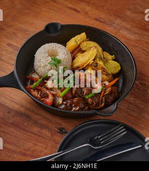 Peruanisches Essen - Lomo saltado - Rinderfilet mit lila Zwiebeln, gelber Paprika, Tomaten in einer schwarzen Pfanne mit pommes frites und Reis serviert. Draufsicht Stockfoto
