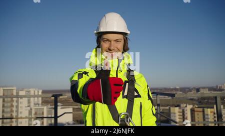 Industriekletterer in weißem Helm zeigt Daumen nach oben und lächelt in die Kamera. Arbeiter im grünen Overall steht auf dem Dach gegen die Stadt Stockfoto