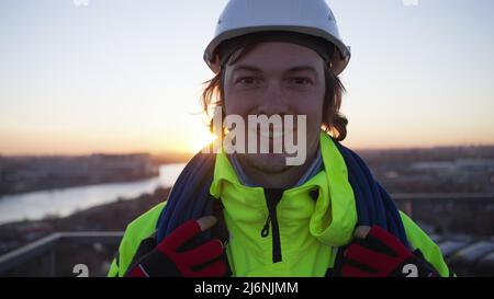 Der junge Industriekletterer in weißem Helm steht bei Sonnenuntergang auf dem Dach. Kletterer mit einem Seil auf den Schultern lächelt in Nahaufnahme in die Kamera Stockfoto