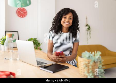 Lächelnde Frau, die ein Smartphone benutzt, während sie zu Hause am Desktop arbeitet. Stockfoto
