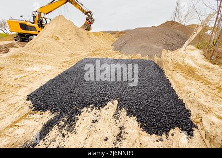 Maschine entleerte heißen und frischen Asphalt in den Sand, in dem sich die Abdrücke der Räder befinden. Bagger gräbt im Hintergrund. Stockfoto