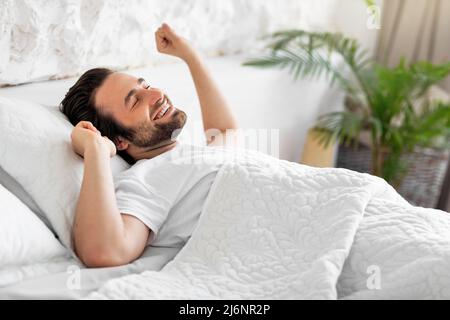 Seitenansicht eines lächelnden jungen Mannes, der sich im Bett streckte Stockfoto