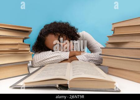 Erschöpfte afroamerikanische Studentin, die auf dem Schreibtisch zwischen Bücherstapeln schläft, während sie sich auf die Prüfung vorbereitet Stockfoto