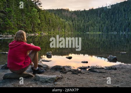 Eine kaukasische Frau mit blonden Haaren und einer roten Jacke, die am Ufer des Sees Certovo jezero im Böhmerwald, Tschechische Republik, sitzt. Nach Sonnenuntergang. Stockfoto