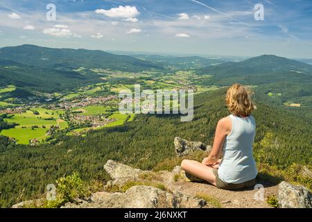 Eine kaukasische Frau, die auf dem Osser-Berg sitzt und die Landschaft von Lamer Winkel mit dem Städtchen Lam überblickt. Bayerischer Wald, Kreis Cham, Oberer