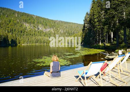 Eine kaukasische Frau sitzt auf der Promenade des schönen Sees großer Arbersee im Bayerischen Wald. Einige Liegestühle vorne. Oberpfalz, Bava Stockfoto