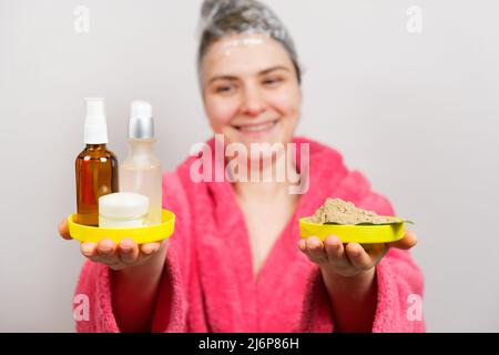 Eine Frau mit einem Film auf dem Kopf macht eine Haarmaske und hält natürliche und unnatürliche Kosmetik in den Händen Stockfoto