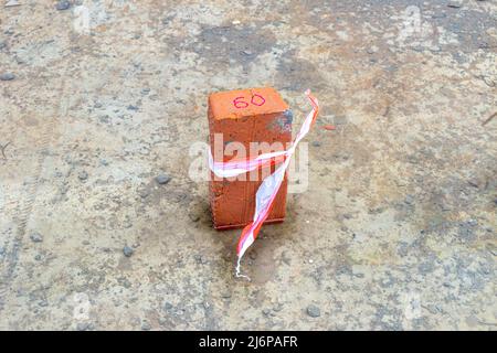 Ein Ziegel mit der roten Aufschrift 60 ist mit einem Signalband gebunden und steht in einer Wasserpfütze auf einer Betonoberfläche, selektiver Fokus Stockfoto