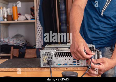 Nicht erkennbarer junger, brünettiger Mann in einem Schlafzimmer, der Desktop-Computer installiert und Kabel und Komponenten miteinander verbindet. Stockfoto